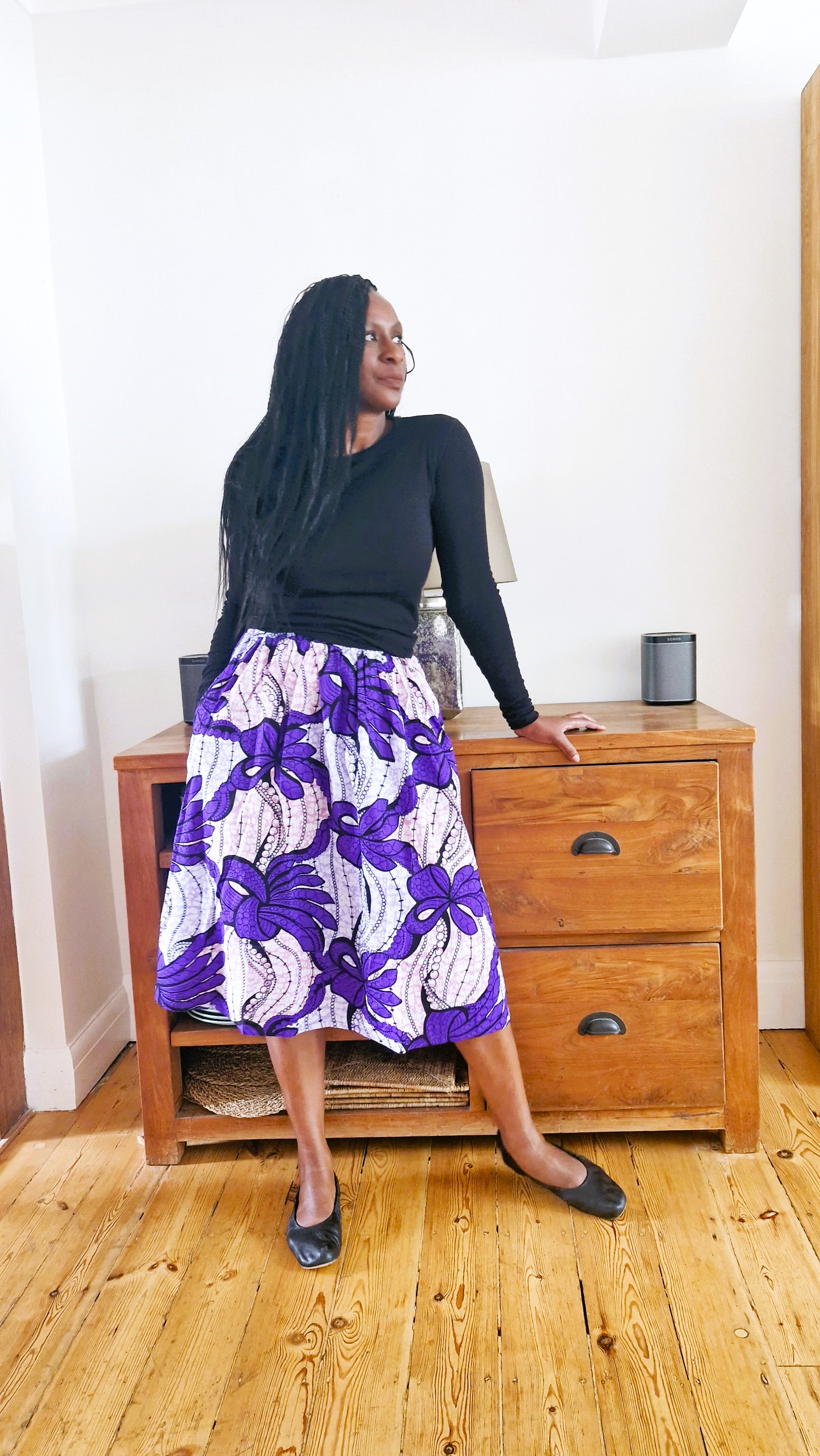 Woman wearing a black top and purple floral skirt standing in a room with wooden furniture.