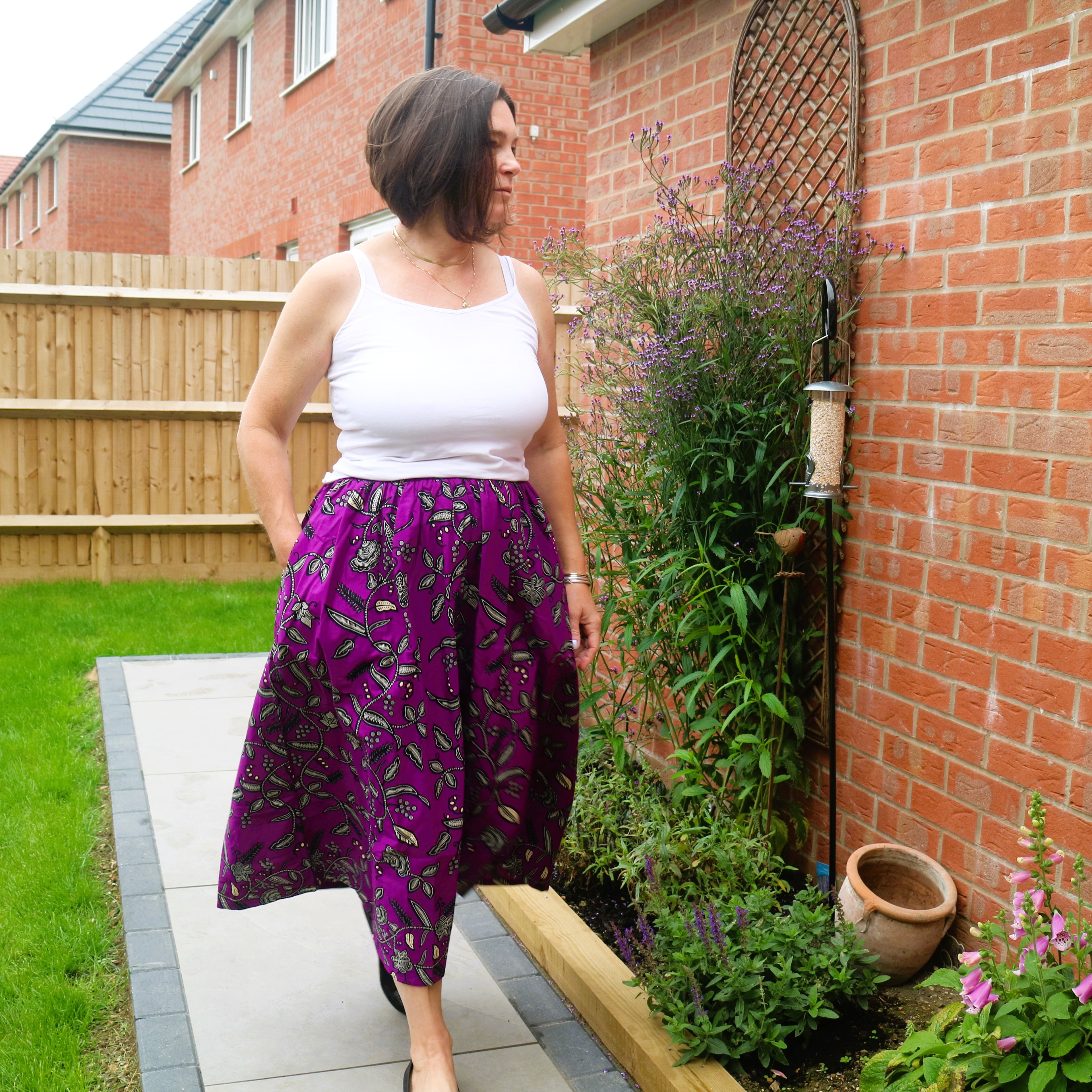 Woman in a white top and purple patterned pants standing in a garden.