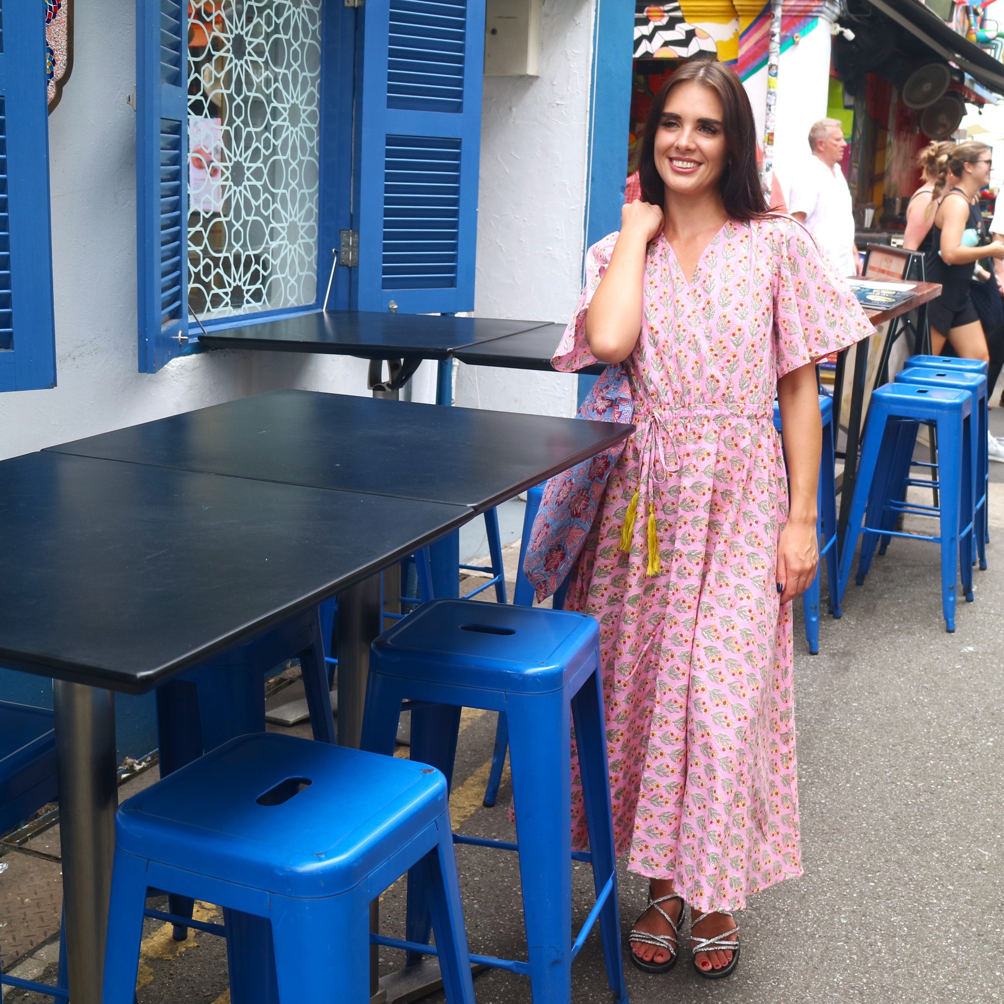 Woman in a pink dress standing next to blue tables and stools in an outdoor setting.