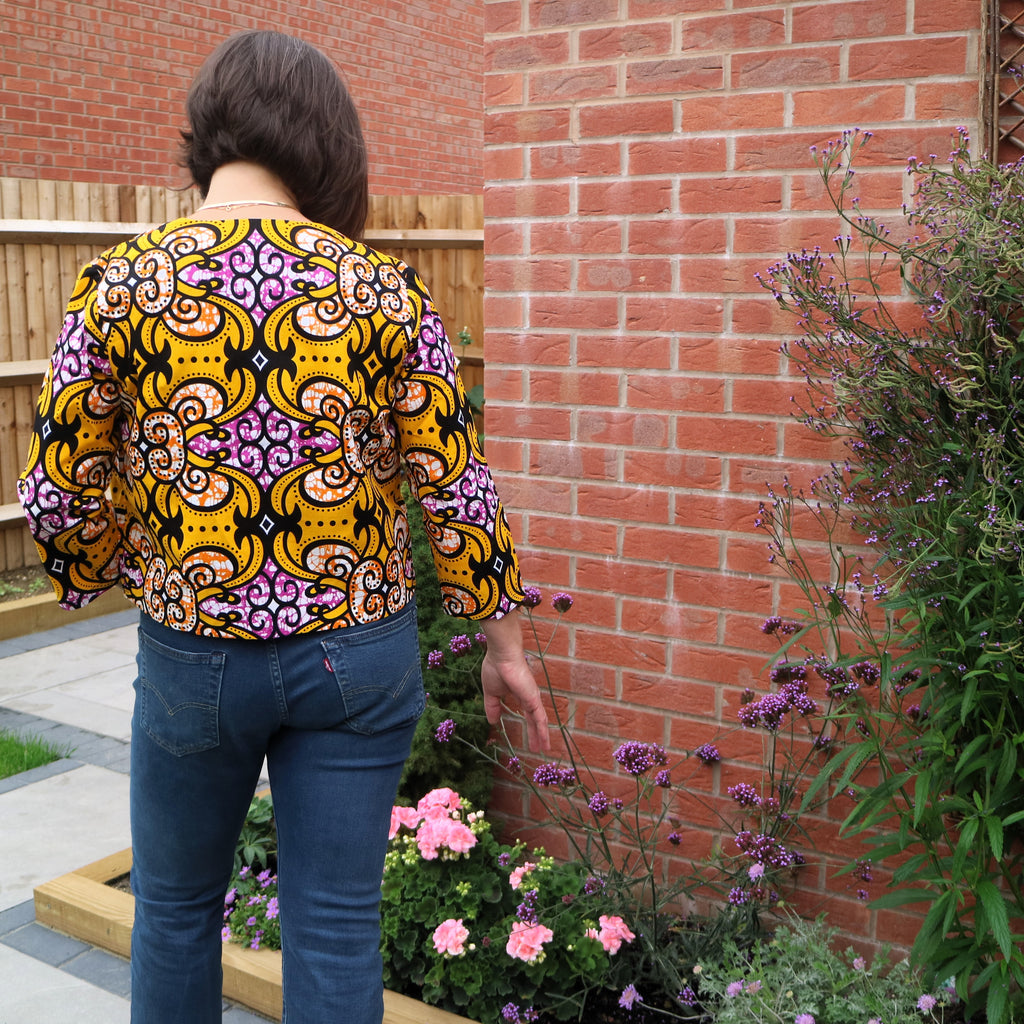 Person wearing a colorful patterned shirt standing in front of a brick wall with plants.