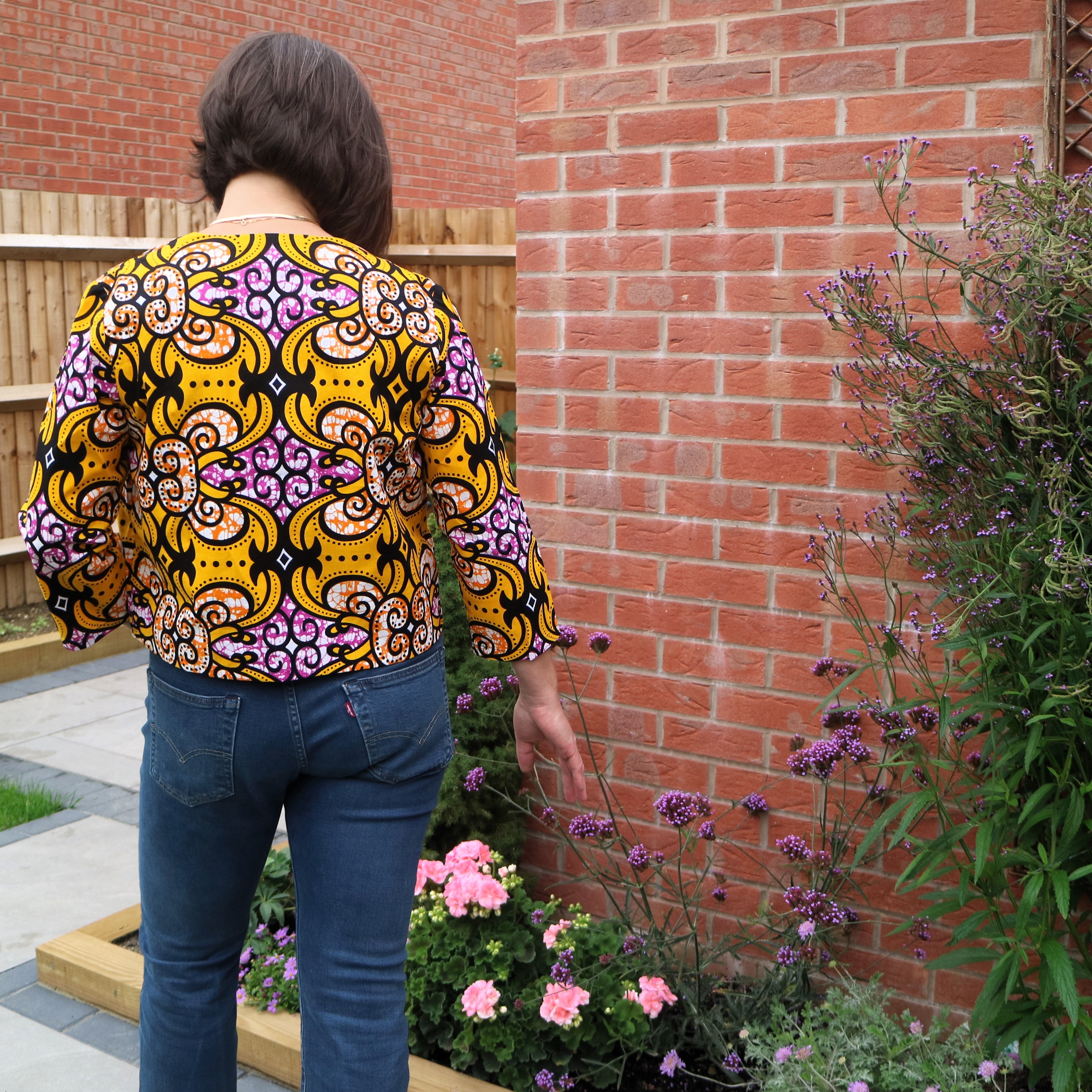 Person wearing a colorful patterned shirt standing in front of a brick wall with plants.